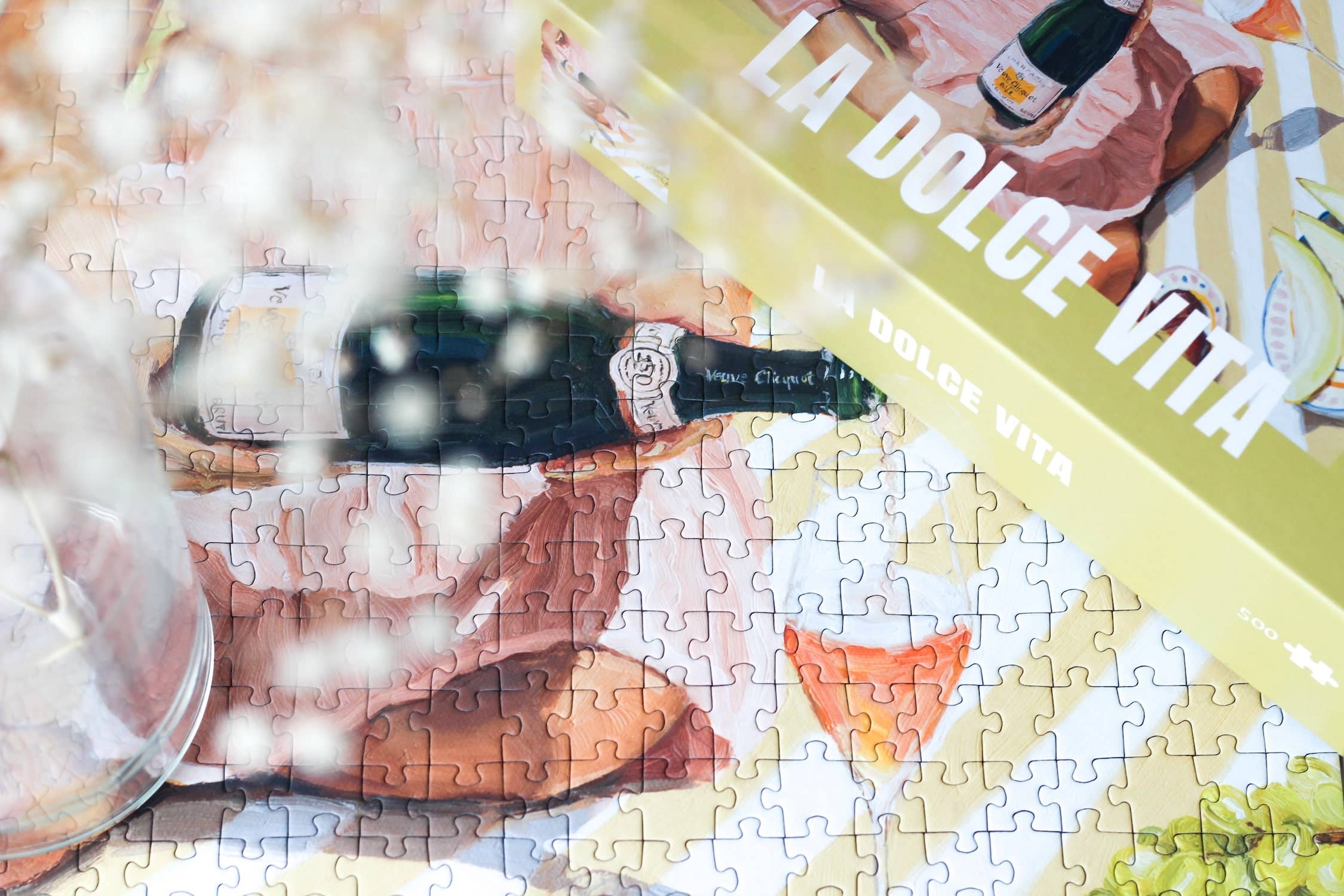 La Dolce Vita jigsaw puzzle box next to a close up section of the completed puzzle featuring a bottle of wine and a glass.